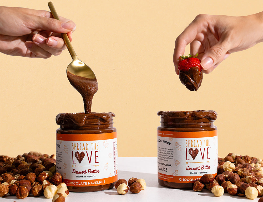 One hand grabbing a spoonful of Chocolate Hazelnut Dessert Butter out of the jar and one hand dipping a strawberry in Chocolate Hazelnut Dessert Butter jar