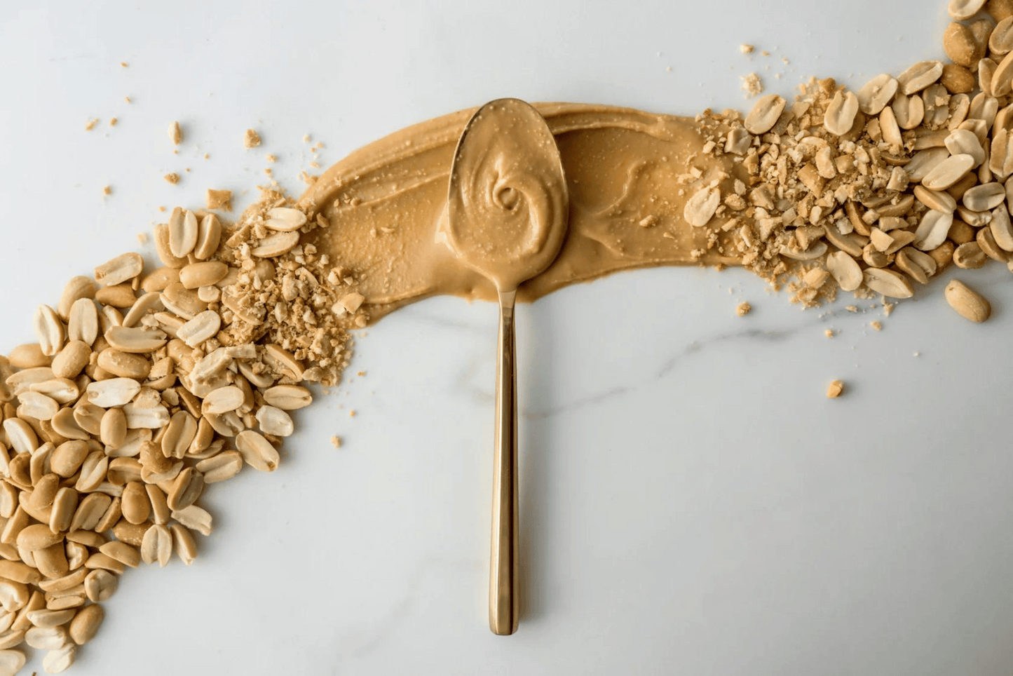 Photo of peanuts and peanut butter spread across counter with spoonful of Peanut Butter in the middle.