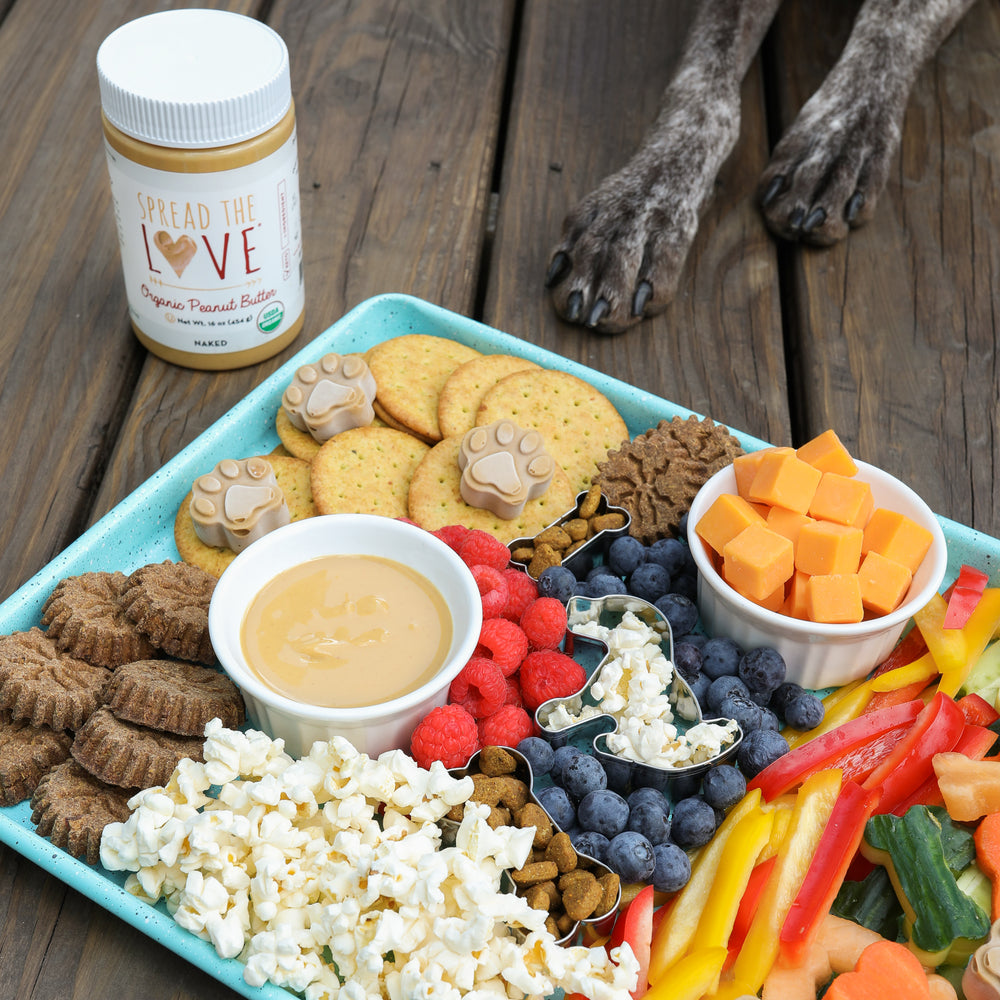 Dog paws and Spread The Love Peanut Butter Jar behind a tray with popcorn, blueberries, dog treats, and veggies