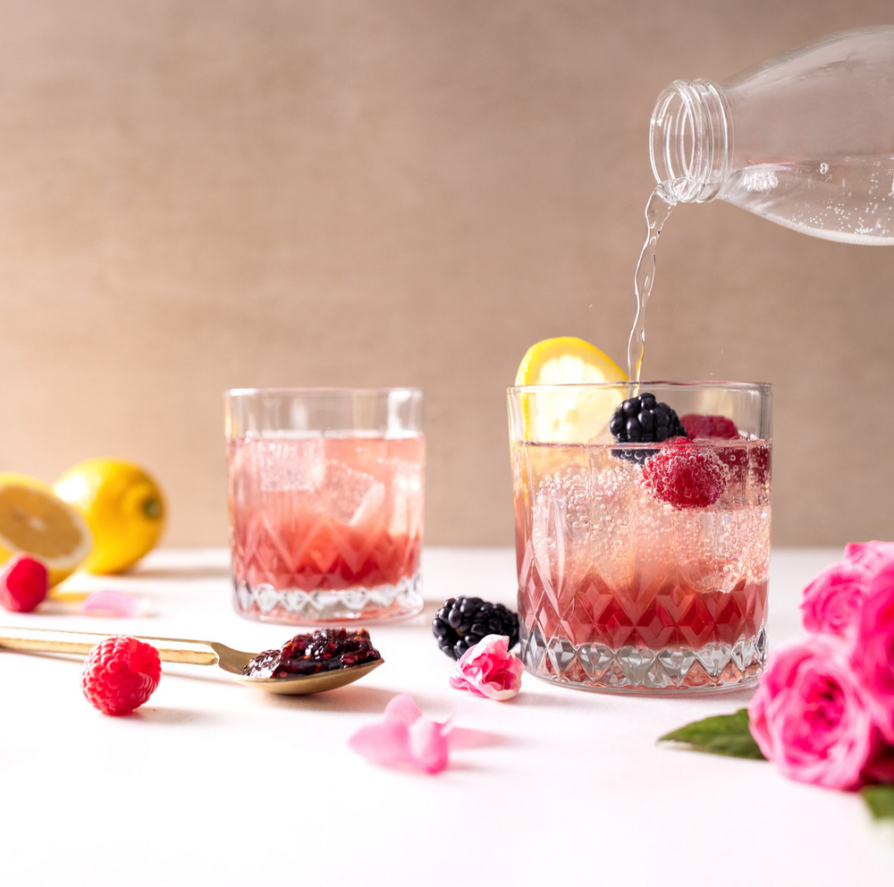 Two glasses of Berry Rose Jam Mocktail surrounded by berries, lemons, and spoonful of jam