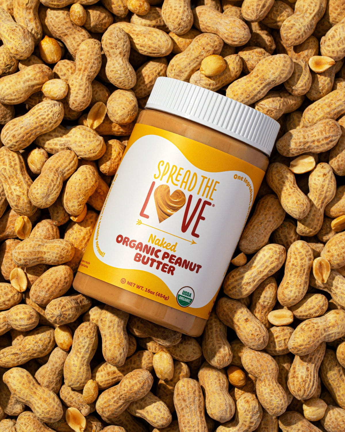Jar of Spread The Love NAKED Organic Peanut Butter surrounded by whole and shelled peanuts, showing the USDA Organic seal and 