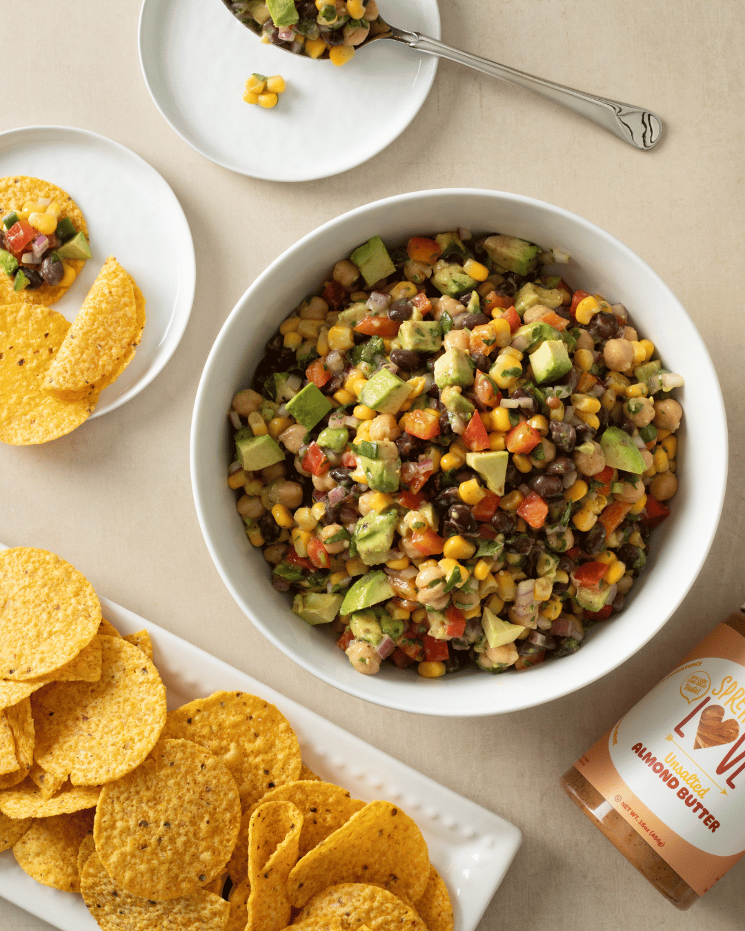 Overhead view of almond butter bean salad in a white bowl surrounded by tortilla chips, serving plates, and Spread The Love UNSALTED Almond Butter