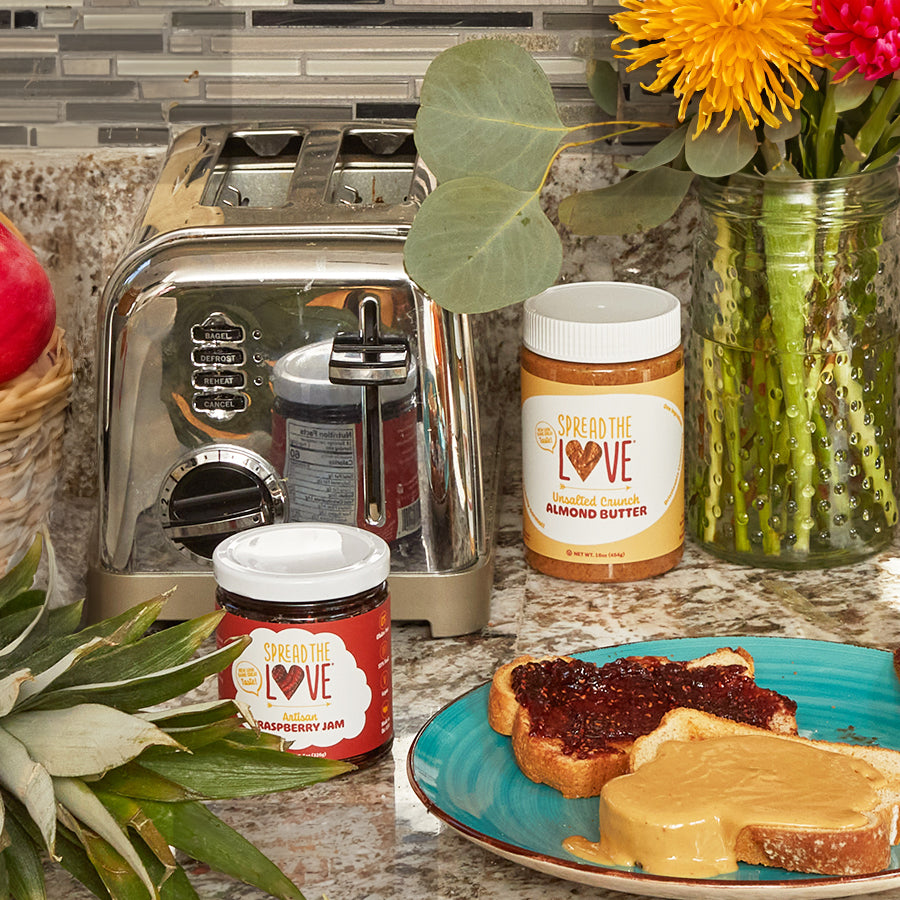 Nut butter and jam on the kitchen counter with fruits around as well as peanut butter and jam sandwich