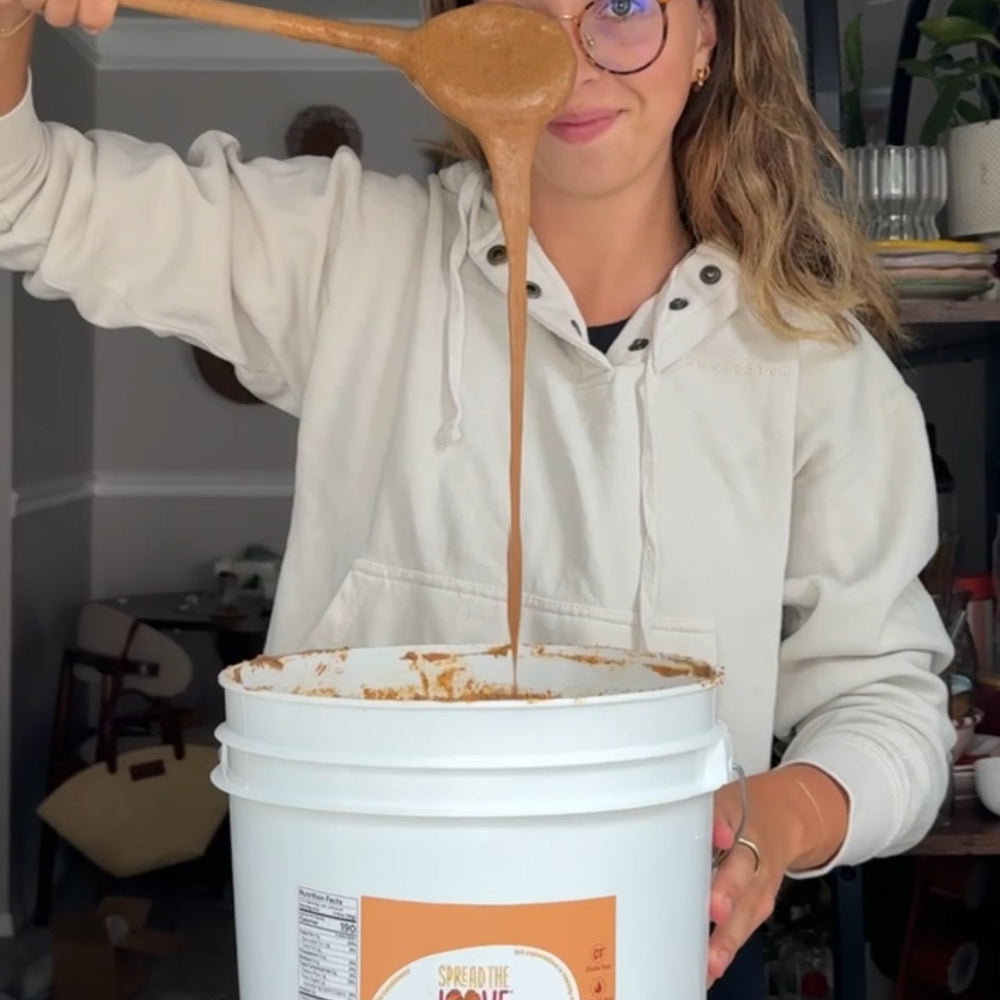 Person holding a wooden spoon with a brown liquid over a white bucket labeled 'Spread the Love'.
