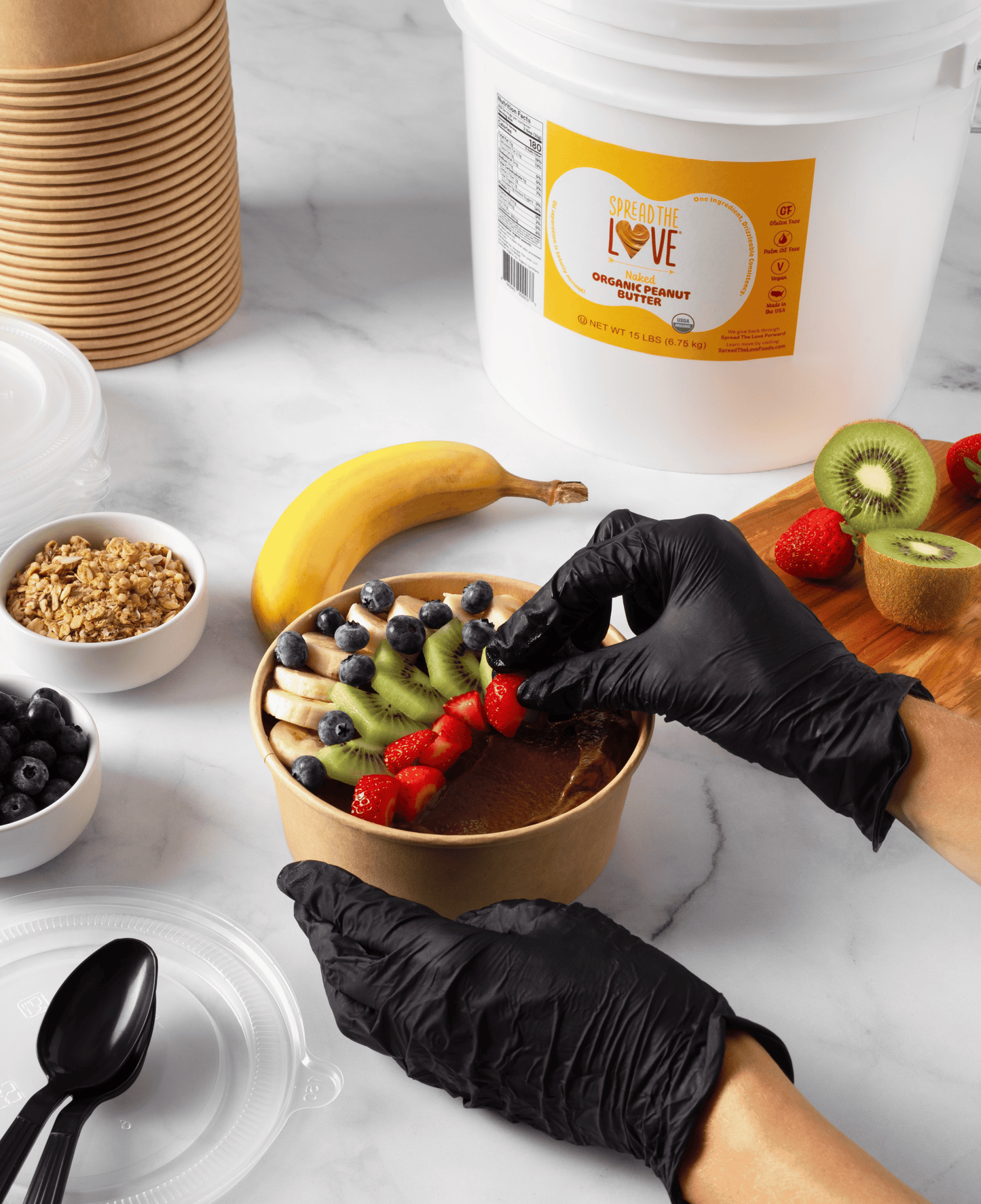 Person preparing smoothie bowl next to Spread The Love Peanut Butter Pail