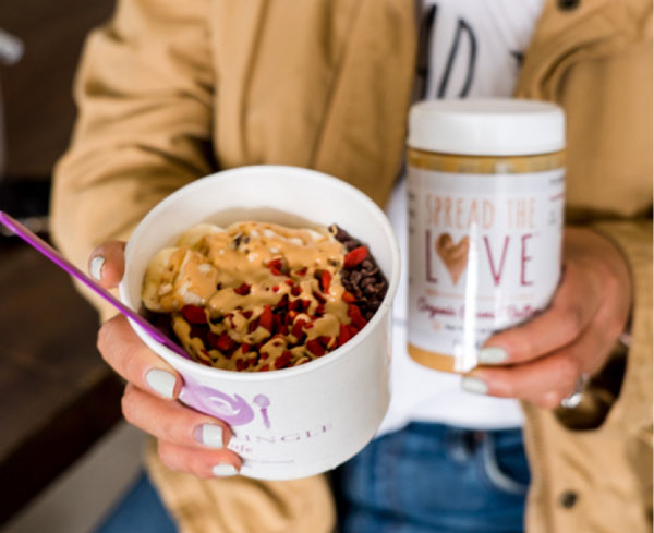 Person holding acai bowl with peanut butter drizzle and Spread The Love Peanut Butter jar
