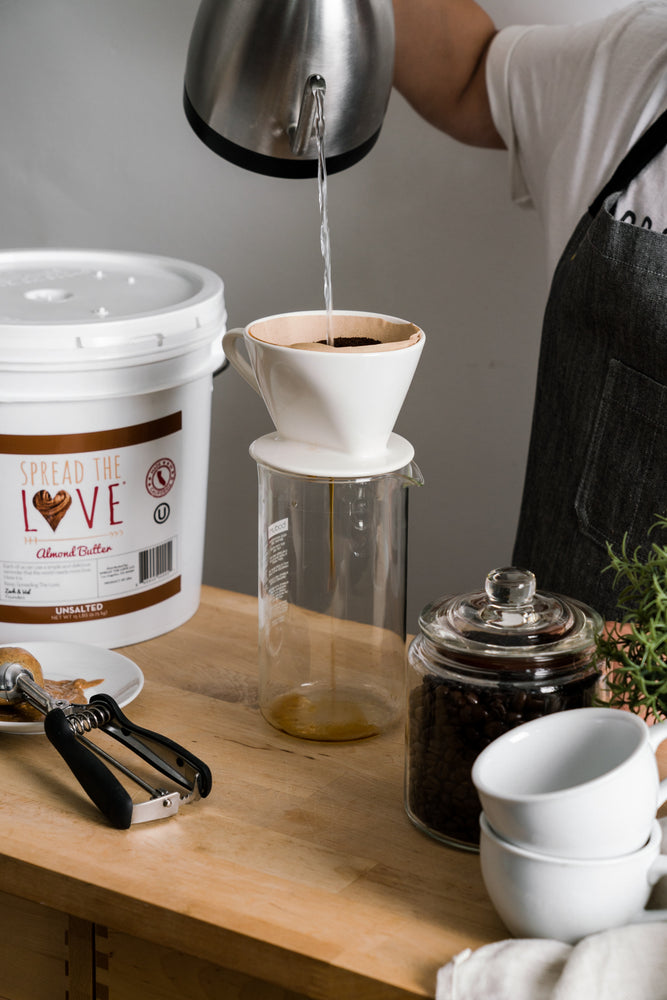 pread The Love Unsalted Almond Butter 40 Pound Pail on a table with a coffee maker, coffee beans, and scoop.